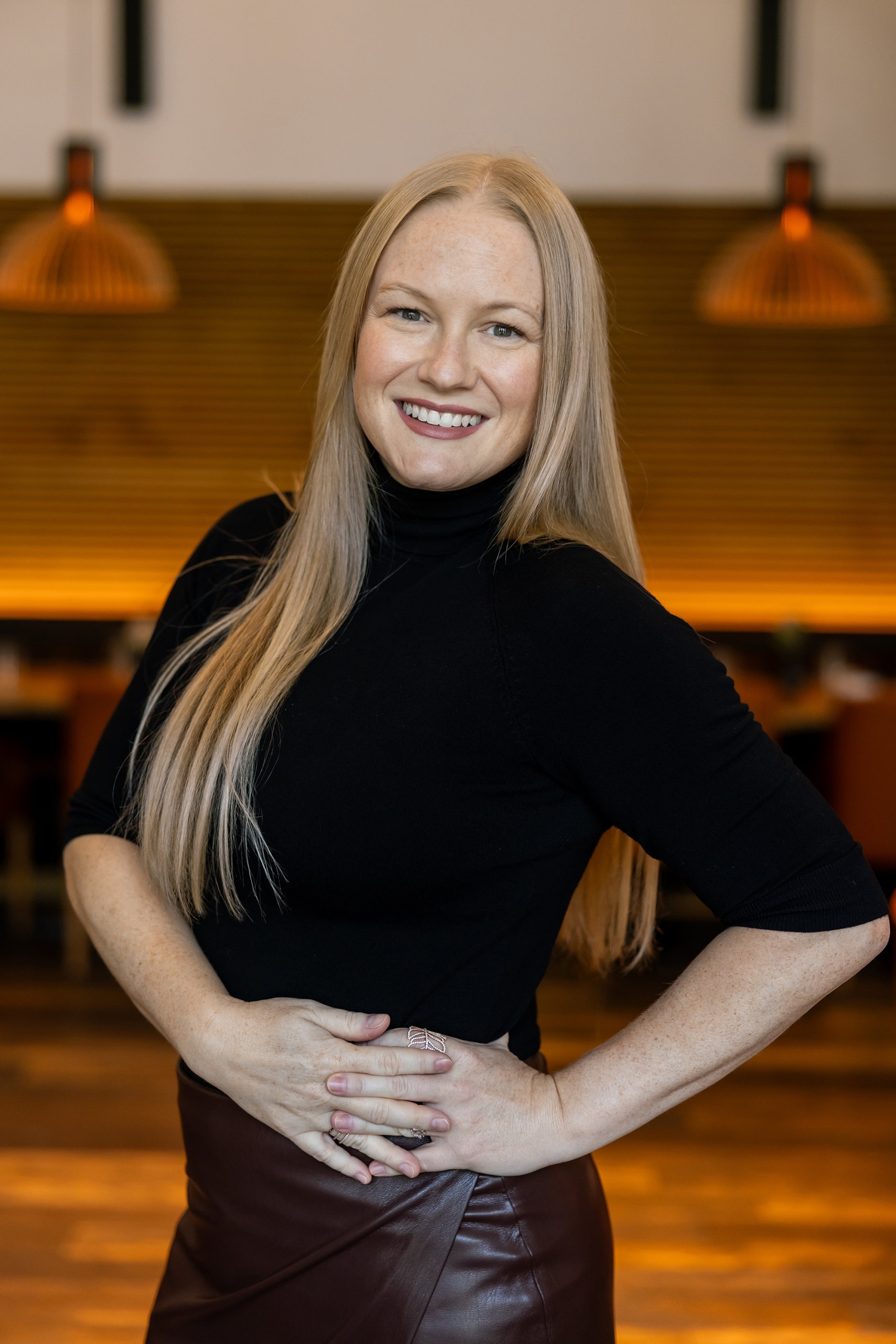 A smiling woman with blonde hair, wearing a black turtleneck, posing in a softly lit restaurant or lounge.