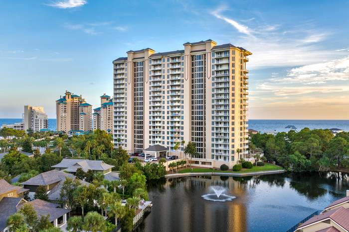 A tall condominium building with a swimming pool at its base, surrounded by other buildings.