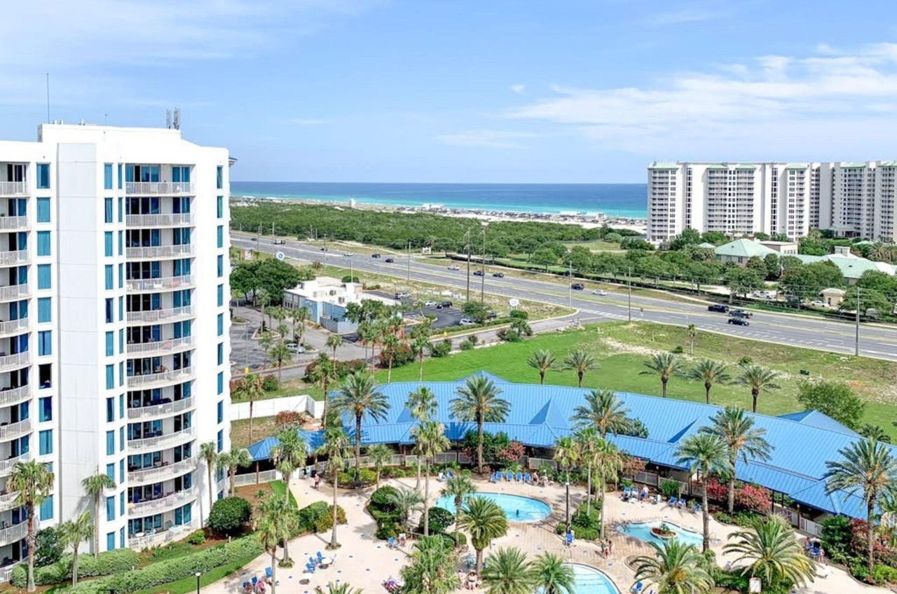 An aerial view of a coastal resort with a tall hotel building overlooking a white-sand beach and the ocean.