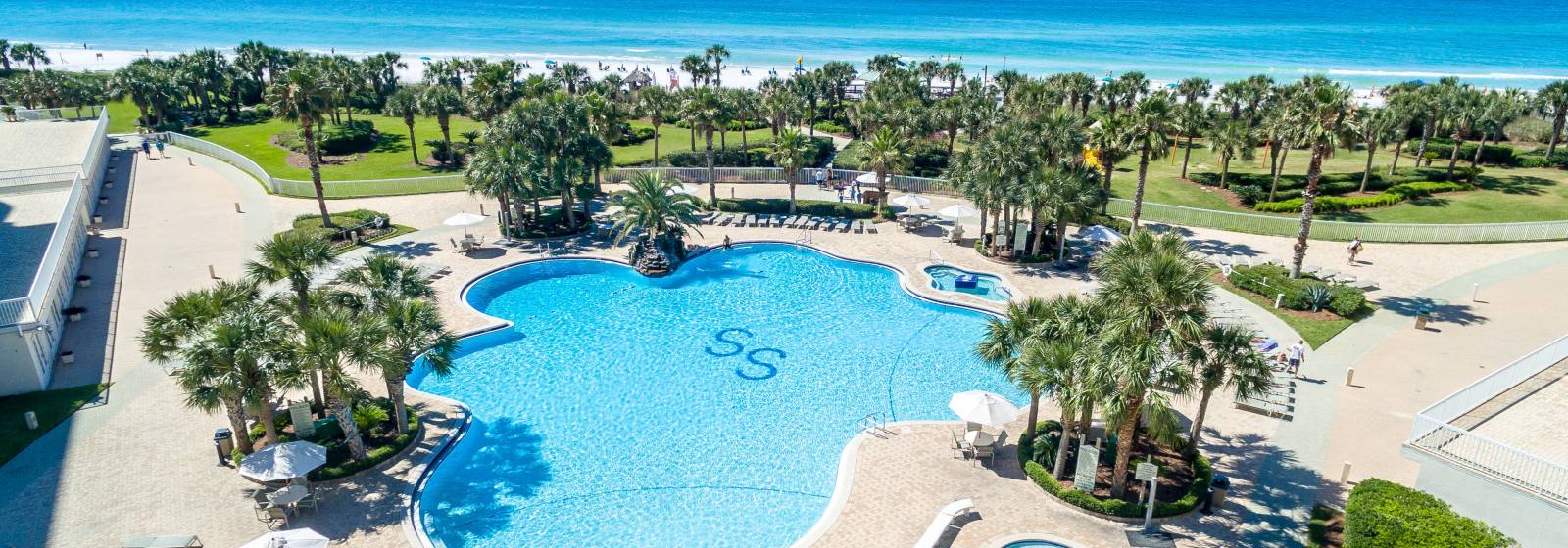 A large, resort-style swimming pool with sparkling blue water, surrounded by palm trees and overlooking the ocean.