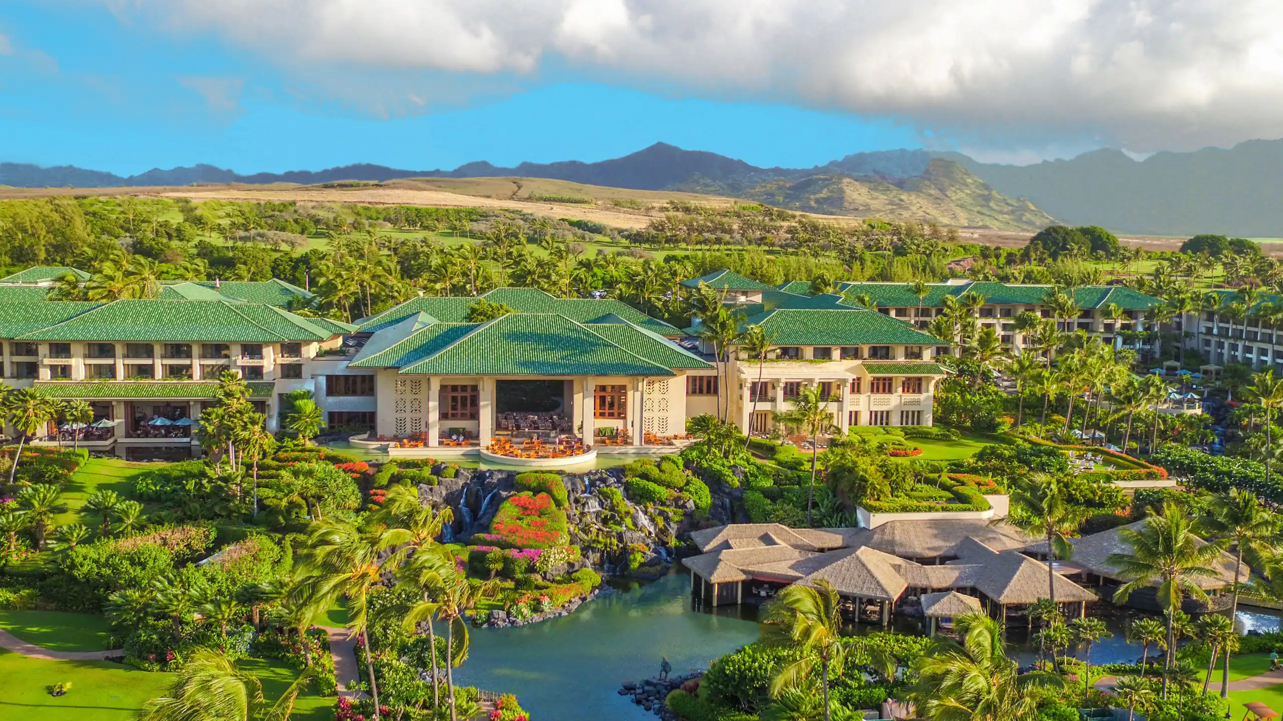 A view of a resort villa with a green tiled roof, surrounded by lush landscaping with mountains in the background.